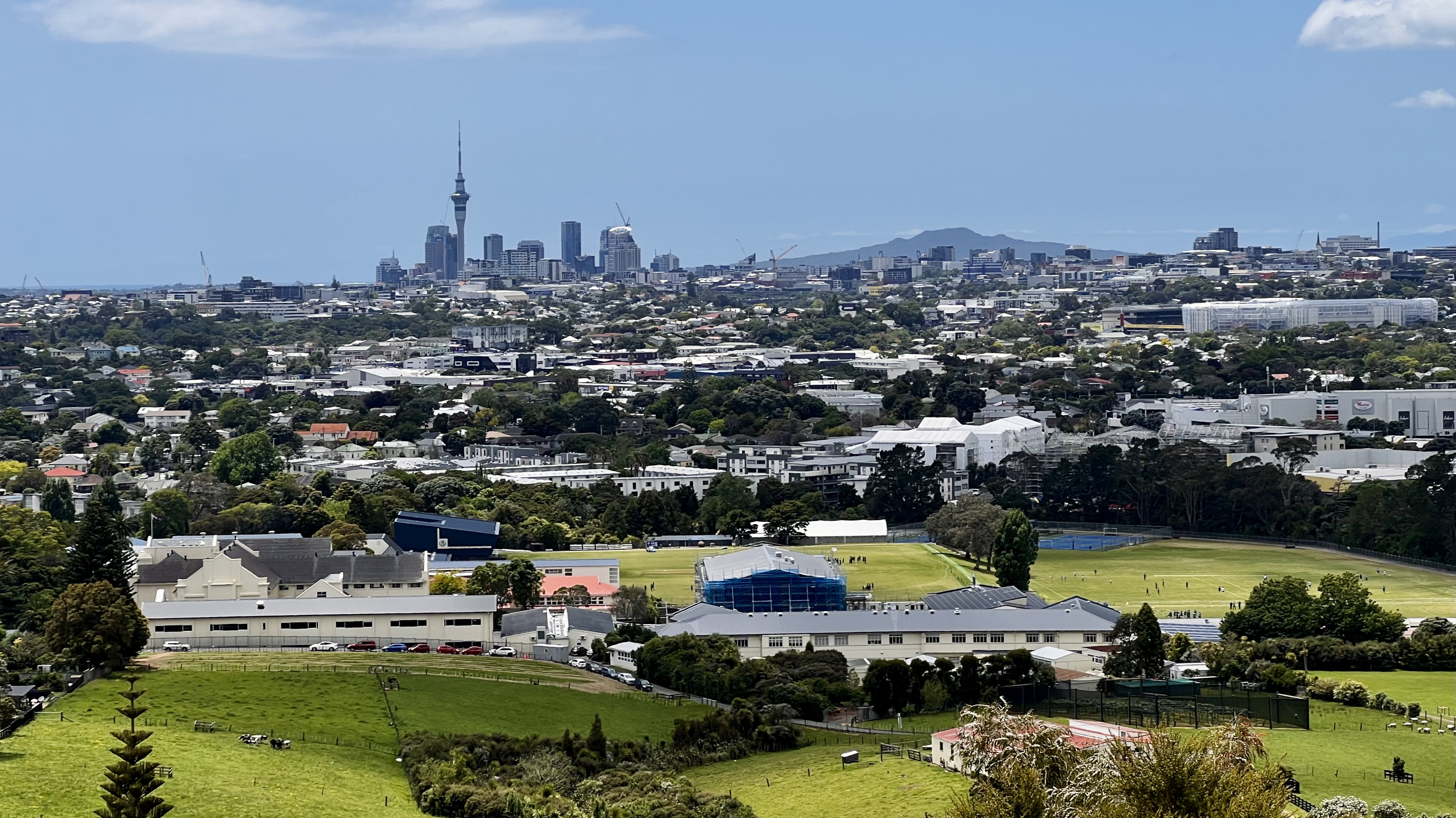Image of the Auckland skyline. Grassy fields in the foreground, neighborhoods in the middle, and tall skyscrapers on the horizon.
